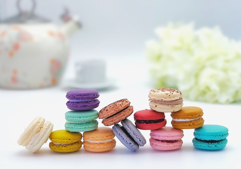 May include: A row of colorful macarons, including purple, teal, yellow, orange, pink, red, brown, and blue, arranged in a semi-circle on a white surface.