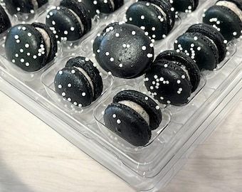 May include: A clear plastic container filled with black macarons with white sprinkles. The macarons are arranged in rows and are all the same size.