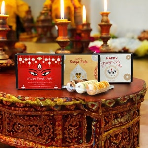 May include: Two gift boxes with the text "Happy Durga Puja" and an image of the Hindu goddess Durga. The boxes are on a wooden table with lit candles in the background. The boxes are filled with small, round, colorful cookies.