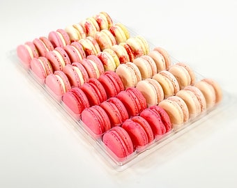 May include: A clear plastic container filled with pink, white, and light pink macarons. The macarons are arranged in rows, with the pink macarons at the bottom, the white macarons in the middle, and the light pink macarons at the top.