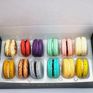 May include: A box of twelve assorted macarons. The macarons are arranged in two rows of six. The macarons are in various colors, including pink, yellow, green, blue, purple, and brown. The box is black with a gold label that says "You to enjoy your moment".