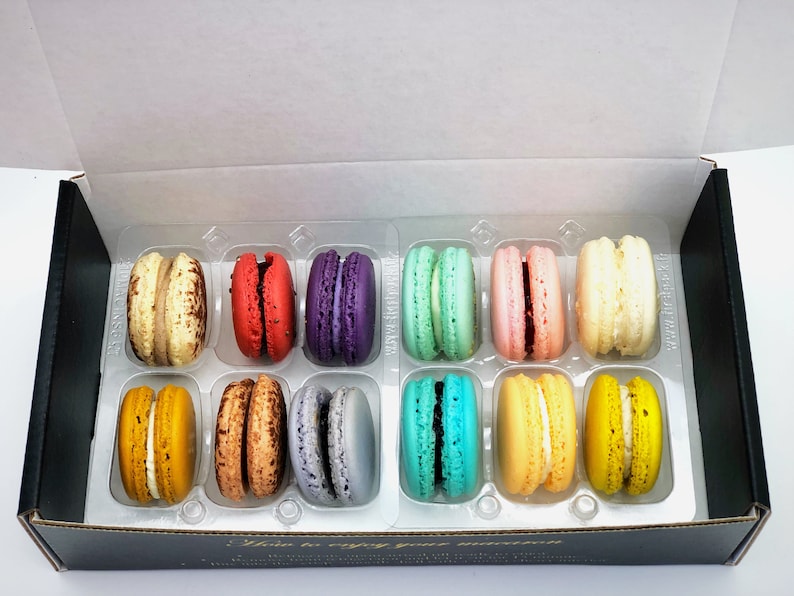 May include: A box of twelve assorted macarons. The macarons are arranged in two rows of six. The macarons are in various colors, including pink, yellow, green, blue, purple, and brown. The box is black with a gold label that says "You to enjoy your moment".
