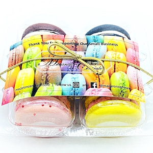 May include: A clear plastic box with a gold ribbon tied around it. The box contains six colorful macarons. The box has a label that says "Thank You for supporting Our Small Business Macaron Centrale macaroncentrale.com follow us @macaroncentrale".