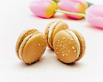 Può includere: Tre macarons, due sono marroni con ripieno bianco e granella bianca, uno è marrone chiaro con ripieno bianco e granella bianca.