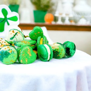 May include: A close-up of green and gold macarons with white frosting. The macarons are arranged in a pile on a white tablecloth.