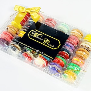 May include: A clear plastic box filled with colorful macarons, tied with a gold ribbon. The box is labeled "Macaron Centrale" with a website and social media handles.