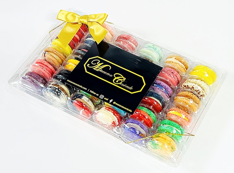 May include: A clear plastic box filled with colorful macarons, tied with a gold ribbon. The box is labeled "Macaron Centrale" with a website and social media handles.