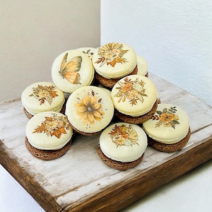 May include: A collection of decorated macarons with a light cream-colored top and a dark brown base. The tops feature various floral and butterfly designs in shades of yellow, orange, and brown. The macarons are arranged on a rustic wooden surface.