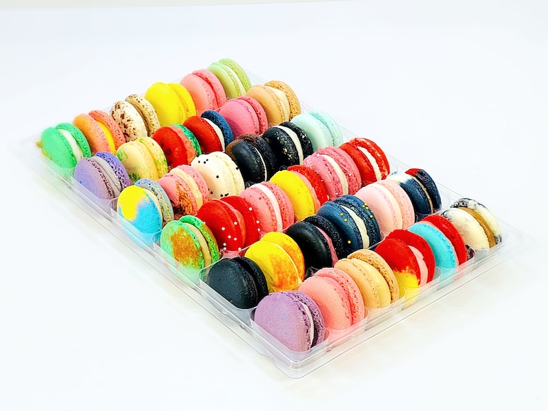 May include: A clear plastic container filled with colorful macarons. The macarons are arranged in rows and are a variety of colors, including pink, blue, green, yellow, and red.
