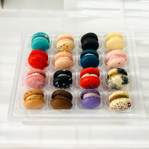 May include: A clear plastic box filled with 16 colorful macarons. The macarons are arranged in a grid pattern and are decorated with sprinkles, chocolate, and other toppings.