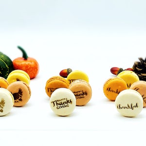 May include: Thanksgiving-themed macarons in various colors, including yellow, orange, and tan. Some macarons feature turkey and text designs. Gourds, acorns, and fall foliage are arranged around the macarons.