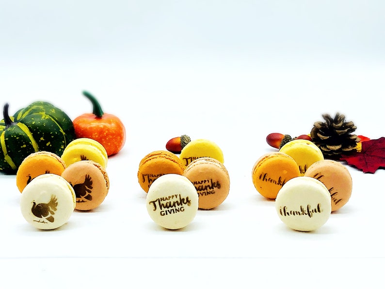 May include: Thanksgiving-themed macarons in various colors, including yellow, orange, and tan. Some macarons feature turkey and text designs. Gourds, acorns, and fall foliage are arranged around the macarons.