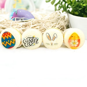 May include: Four Easter-themed macarons with different designs. One macaron has a blue and yellow egg with a zig-zag pattern, another has the words "Happy Easter" in a decorative font, a third has a white bunny with pink ears, and the last has a yellow egg with a pink flower.