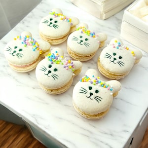 May include: Six white bunny-shaped macarons with colorful sprinkles on top. The macarons have black painted eyes and whiskers.