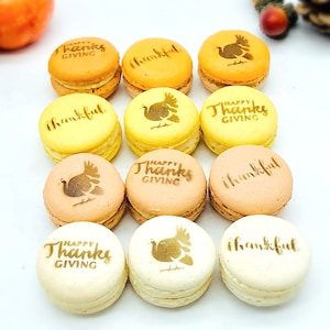 May include: A dozen colorful macarons in shades of yellow, orange, and cream. Each macaron is decorated with the words "Happy Thanksgiving" or "thankful", and a turkey graphic. The macarons are arranged on a white surface.
