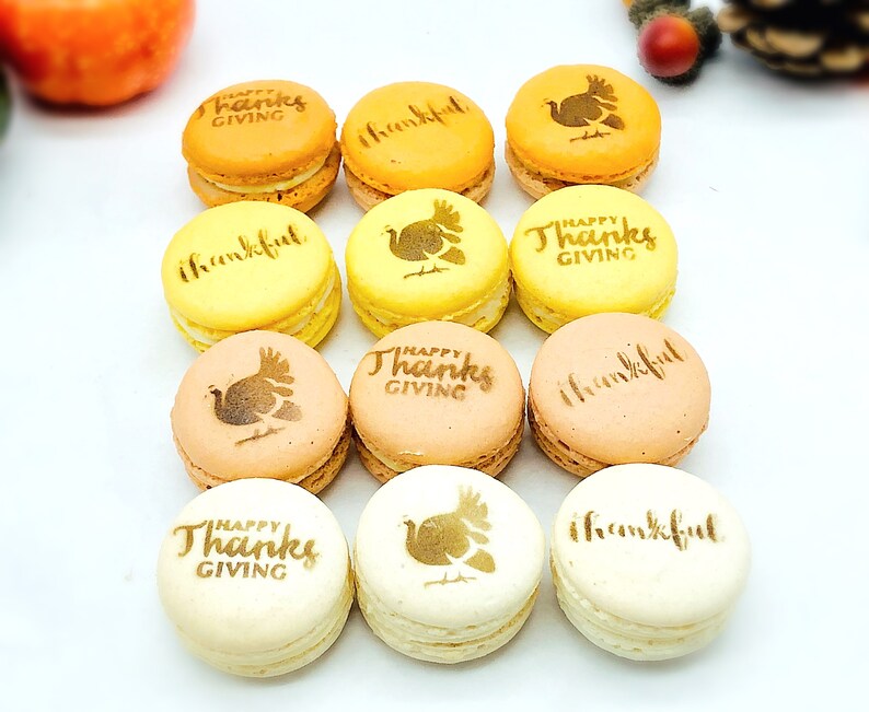 May include: A dozen colorful macarons in shades of yellow, orange, and cream. Each macaron is decorated with the words "Happy Thanksgiving" or "thankful", and a turkey graphic. The macarons are arranged on a white surface.