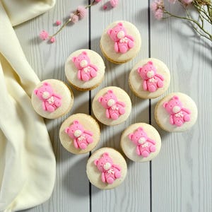 May include: A top-down shot of ten macarons, each topped with a pink teddy bear decoration. The macarons are arranged on a light gray wooden surface, with a cup of coffee and pink flowers in the background.