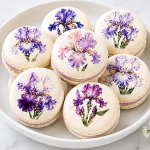 May include: A collection of six round, cream-colored macarons arranged in a white bowl. Each macaron features a detailed, printed illustration of a purple and white iris flower on its top surface, with a visible light purple filling between the shells.