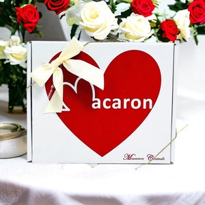 May include: White gift box with a red heart and the word "macaroon" printed on it. The box is tied with a cream-colored ribbon and bow. The text "Macaron Centrale" is printed on the bottom right corner of the box.