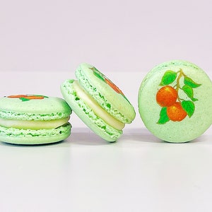 May include: Three green macarons with white filling and orange fruit designs. The macarons are arranged in a row on a white surface.