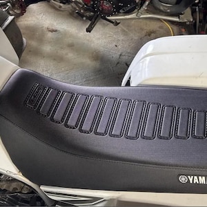 May include: A black and white Yamaha ATV seat with a black, padded seat and a black and white "YAMAHA" logo. The seat has a black, textured surface with a series of rectangular, stitched panels. The seat also has a black "LCM" logo on the right side.