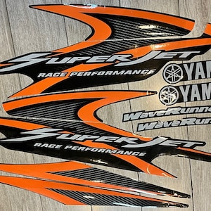 Puede incluir: Un conjunto de calcomanías negras y naranjas con las palabras "Super Jet" y "Race Performance" en blanco. Las calcomanías también presentan el logotipo de Yamaha y las palabras "WaveRunner".