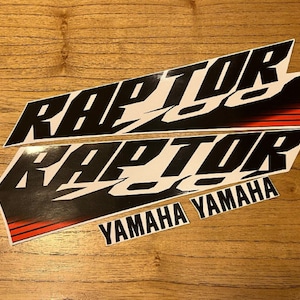 以下が含まれることがあります： スタイリッシュなフォントで「RAPTOR」という単語が書かれた、黒と白の2つのデカール。 下のデカールには、黒文字で「YAMAHA」という単語も含まれています。