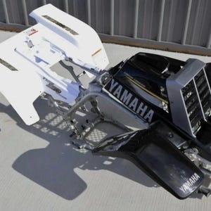 Peut inclure: Un kit de carrosserie pour VTT Yamaha comprenant une section de garde-boue arrière blanche avec des autocollants noirs et un assemblage de garde-boue avant noir avec un cache-radiateur argenté. Le logo Yamaha blanc est visible sur les panneaux.