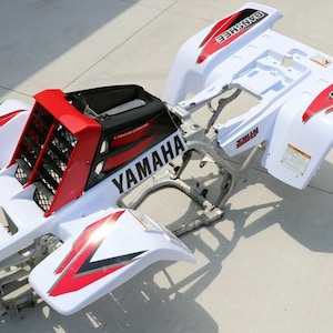 Peut inclure: Cadre de quad Yamaha Banshee blanc avec des graphismes rouges et noirs. Le cadre est démonté et les garde-boue avant et arrière sont retirés. Le cadre est en métal et les garde-boue sont en plastique.