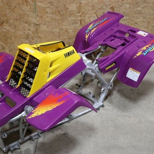 Peut inclure: Un cadre de quad Yamaha Banshee 350 avec des panneaux de carrosserie en plastique violet et jaune. Le cadre est argenté et les panneaux ont des flammes et le mot "Banshee" imprimé dessus.
