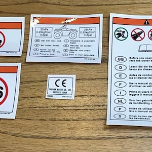 Op de afbeelding: Een set van acht oranje en witte waarschuwingstickers met zwarte tekst en symbolen. De stickers bevatten instructies voor het bedienen van een voertuig in meerdere talen, een CE-markering en een waarschuwingsbord met de tekst "<16".