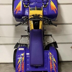 May include: A purple and yellow Yamaha ATV with a black handlebar and a black seat. The ATV has a graphic of a flame on the side with the word "FISTIC" on it.