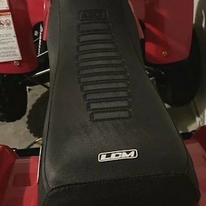 Könnte beinhalten: Schwarzer ATV-Sitzbezug mit strukturiertem Design und dem aufgedruckten Wort "LCM". Der Sitzbezug ist auf einem roten ATV montiert.