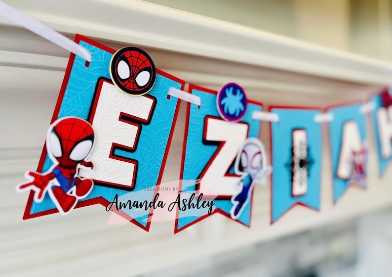 K&ouml;nnte beinhalten: Ein blaues und rotes Banner mit dem Namen "EZIAH" in wei&szlig;en Buchstaben. Jeder Buchstabe hat einen anderen Spiderman-Charakter darauf. Das Banner hat auch den Text "CREATIONS BY Amanda Ashley".