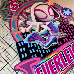 May include: A layered cake topper featuring a Spider-Gwen character design. The topper includes a number 5, a spider web, and a cityscape. The color scheme is primarily pink, teal, and purple, with holographic accents.