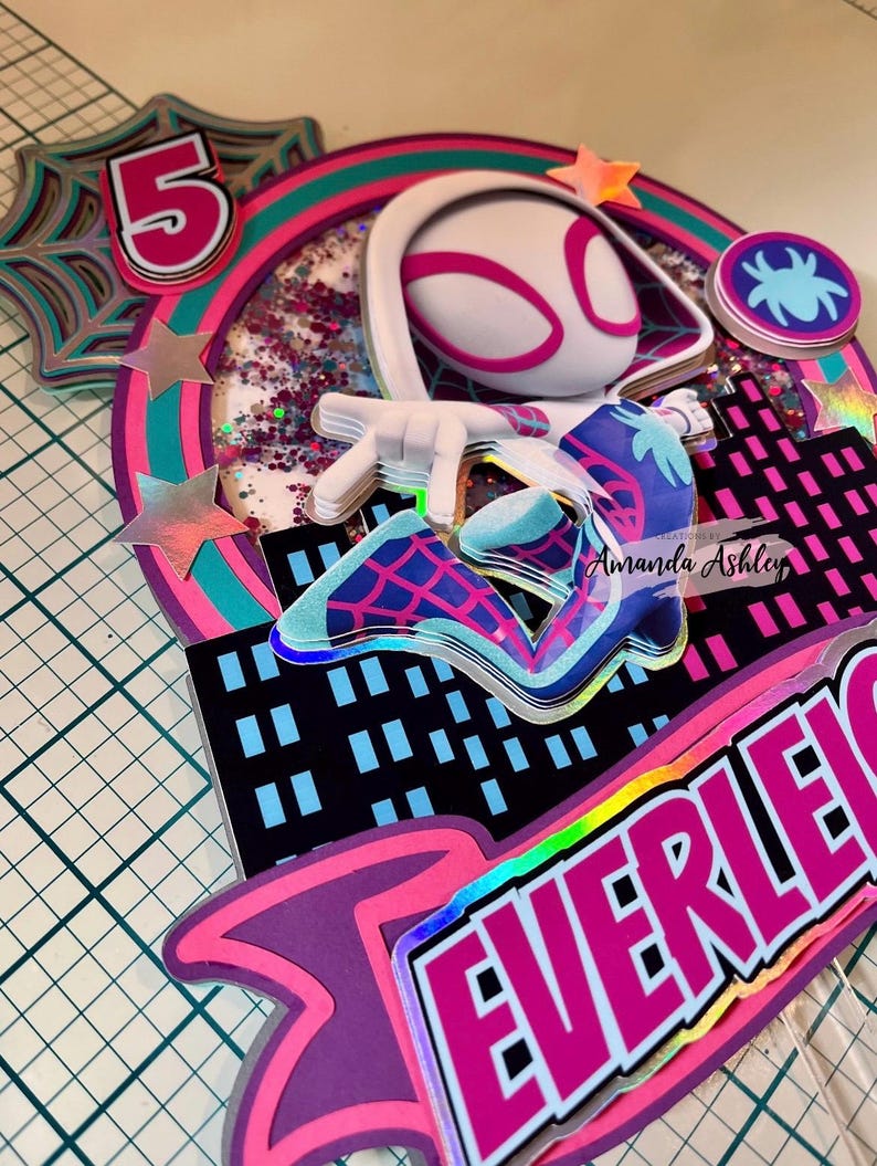 May include: A layered cake topper featuring a Spider-Gwen character design. The topper includes a number 5, a spider web, and a cityscape. The color scheme is primarily pink, teal, and purple, with holographic accents.