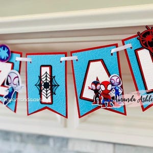 K&ouml;nnte beinhalten: Ein blaues und rotes Banner mit dem Namen "EZIAH" in wei&szlig;en Buchstaben. Jeder Buchstabe hat einen kleinen Spiderman-Charakter darauf. Das Banner ist mit einem Spinnennetz-Muster verziert.