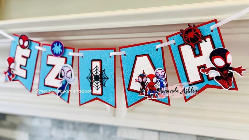 K&ouml;nnte beinhalten: Ein blaues und rotes Banner mit dem Namen "EZIAH" in wei&szlig;en Buchstaben. Jeder Buchstabe hat einen kleinen Spiderman-Charakter darauf. Das Banner ist mit einem Spinnennetz-Muster verziert.