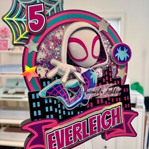 May include: A multi-layered cake topper featuring a Spider-Gwen character with the number 5. The design includes a cityscape, stars, and the name "EVERLEIGH" in a pink and purple banner. The topper has a clear acrylic base.