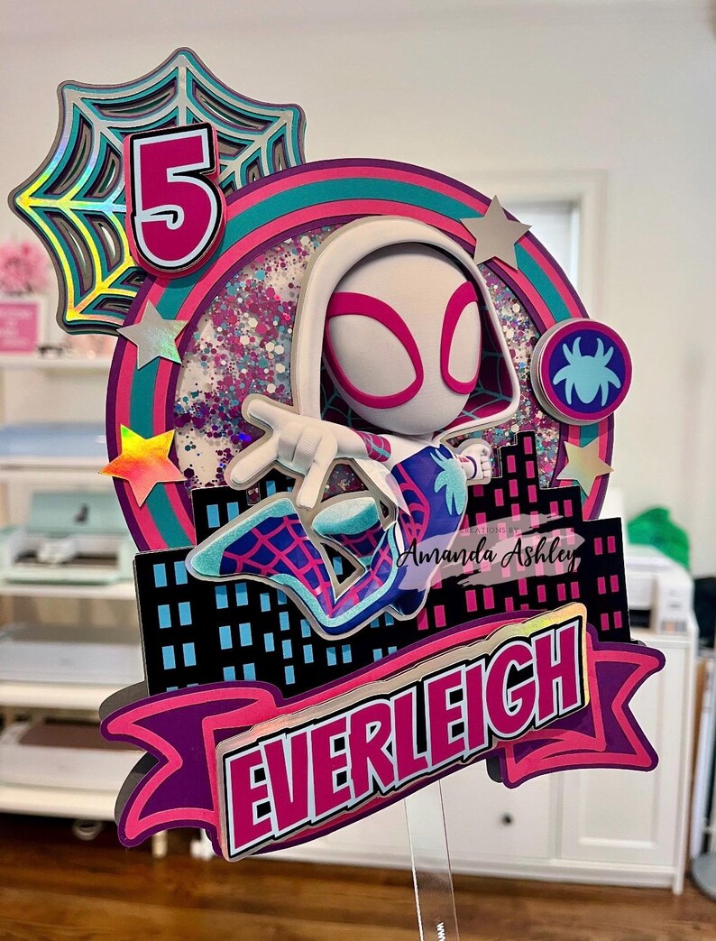 May include: A multi-layered cake topper featuring a Spider-Gwen character with the number 5. The design includes a cityscape, stars, and the name "EVERLEIGH" in a pink and purple banner. The topper has a clear acrylic base.