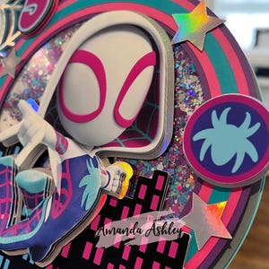 May include: A colorful, layered cake topper featuring a Spider-Gwen character design. The design includes a number 5, a spider web, and a cityscape. The topper has a circular design with glitter and holographic stars. Text reads "Creations by Amanda Ashley."
