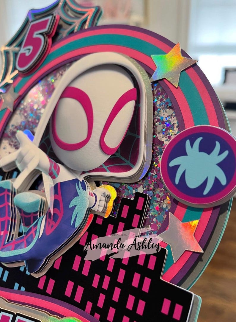 May include: A colorful, layered cake topper featuring a Spider-Gwen character design. The design includes a number 5, a spider web, and a cityscape. The topper has a circular design with glitter and holographic stars. Text reads "Creations by Amanda Ashley."