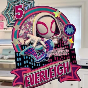 May include: A colorful cake topper featuring a superheroine character with a white and pink mask and costume. The design includes a cityscape, the number 5, and the name "Everleigh". The topper is decorated with glitter and star accents.