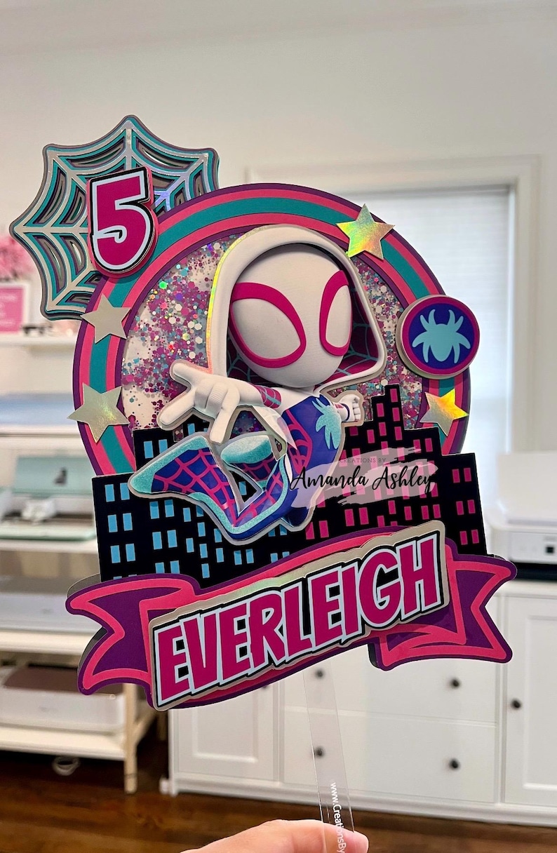 May include: A colorful cake topper featuring a superheroine character with a white and pink mask and costume. The design includes a cityscape, the number 5, and the name "Everleigh". The topper is decorated with glitter and star accents.