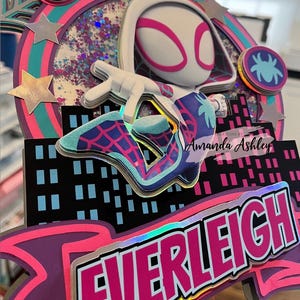 May include: A colorful cake topper featuring a Spider-Gwen character design. The topper includes the name "EVERLEIGH" in a vibrant pink and white font, along with a cityscape silhouette and star accents. The number "5" is also visible.