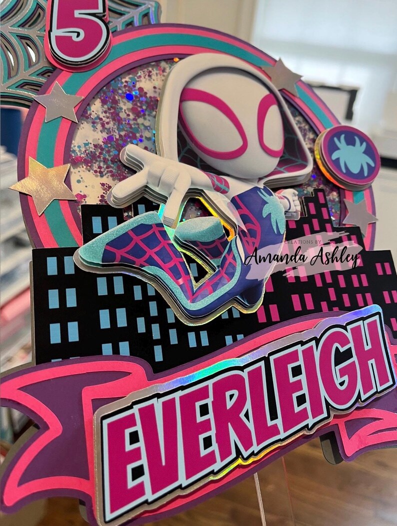 May include: A colorful cake topper featuring a Spider-Gwen character design. The topper includes the name "EVERLEIGH" in a vibrant pink and white font, along with a cityscape silhouette and star accents. The number "5" is also visible.