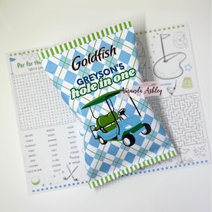 Puede incluir: Una bolsa de galletas Goldfish con temática de golf. La bolsa tiene un diseño de cuadros azules y blancos y una ilustración de carrito de golf. El texto en la bolsa dice "GREYSON'S hole in one". Un juego de búsqueda de palabras y un laberinto están en el fondo.