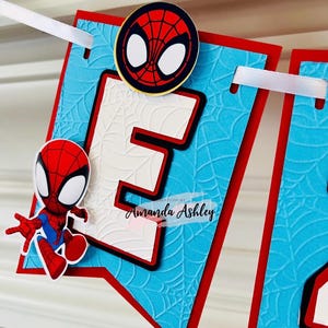 K&ouml;nnte beinhalten: Ein blaues und rotes Banner mit einem wei&szlig;en Buchstaben E und einer Spiderman-Figur. Das Banner hat ein Spinnennetz-Muster und den Text "Creations by Amanda Ashley".