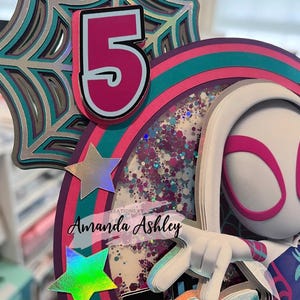 May include: A multi-layered birthday cake topper featuring a number 5 in pink and white, a spider web design, and a character with a white mask and pink accents. The topper includes iridescent stars and the text "Creations by Amanda Ashley."
