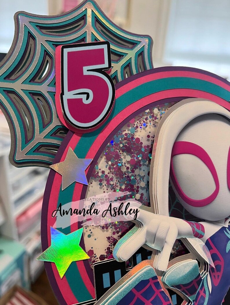 May include: A multi-layered birthday cake topper featuring a number 5 in pink and white, a spider web design, and a character with a white mask and pink accents. The topper includes iridescent stars and the text "Creations by Amanda Ashley."